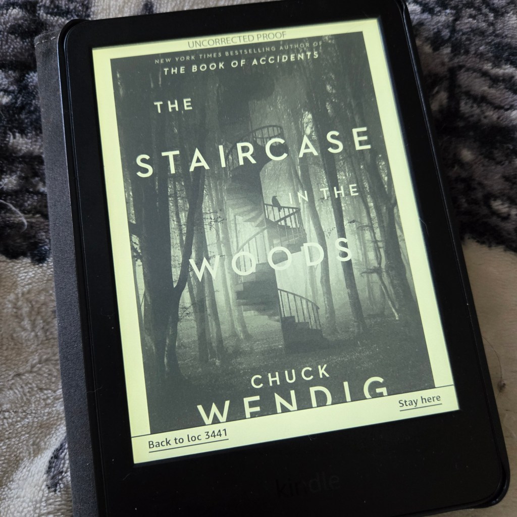 A kindle reader with the cover of The Staircase in the Woods displayed in black and white. The cover shows a spiral staircase in the forest with a bird perched on the railing.